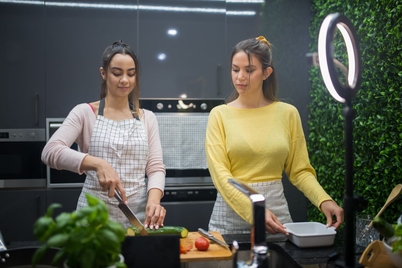 services-04 Two women cooking and preparing food in a modern kitchen with a ring light. Collaborative culinary activity.